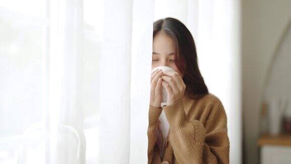 年轻的亚洲妇女站在窗户旁边在家里的客厅拿着杯子亚洲女性早上醒来后喝牛奶或茶