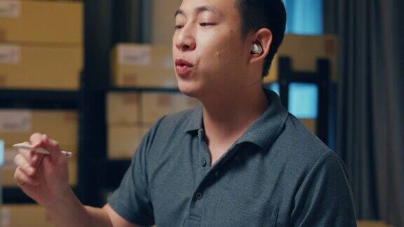 亚洲商人戴着耳机使用数字平板电脑在仓库快递前与客户在线查看库存数据详细信息