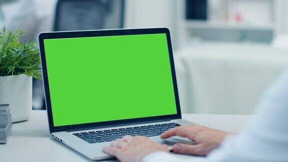 一个医生在电脑上工作的特写镜头绿色的屏幕他在后台的同事办公室是现代和良好的照明