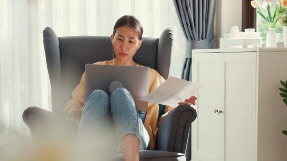 年轻的亚洲女女孩或大学生坐在沙发椅上带着电脑笔记本电脑戴着耳机在家里的客厅里举行文件在线会议展示讨论工作