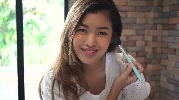 年轻的亚洲微笑女人使用化妆刷和看着相机在家里