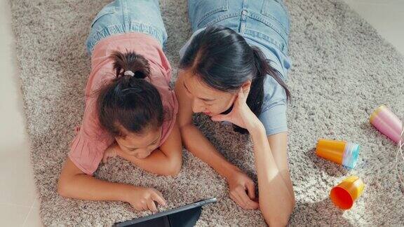 亚洲蹒跚学步的小女孩女儿和妈妈在地毯上看数码平板电脑有乐趣在家里的客厅快乐的时刻