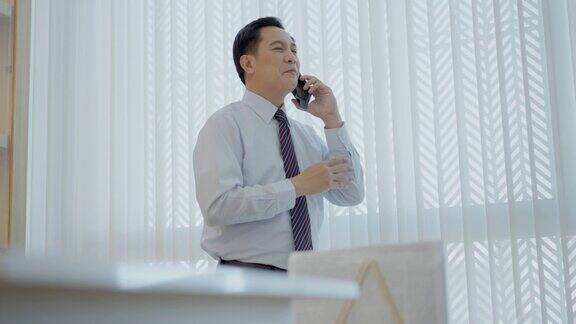 英俊的高级财务顾问站在办公室的大窗户旁一边用手机向尊贵的客户讲解投资计划下一步的商业财务计划年收入丰厚