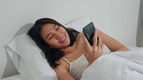 年轻的亚洲女人使用智能手机查看社交媒体早上醒来后躺在床上开心地微笑美丽迷人的西班牙女孩微笑放松在卧室在家里