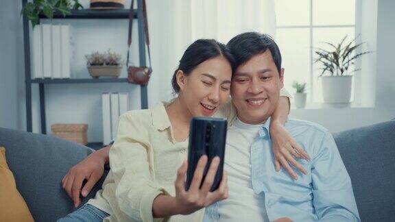 年轻的亚洲夫妇与休闲坐在沙发上使用智能手机自拍在线呼叫挥手手势与家人和朋友在家里的客厅