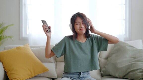 年轻的亚洲女子在家里客厅的沙发上听音乐快乐的亚洲女性使用手机戴着耳机坐在沙发上