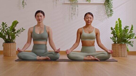一对亚洲年轻女子坐在瑜伽垫上做呼吸练习一起做莲花瑜伽瑜伽冥想两位健康女性在舒适的家中放松舒适健康的生活方式