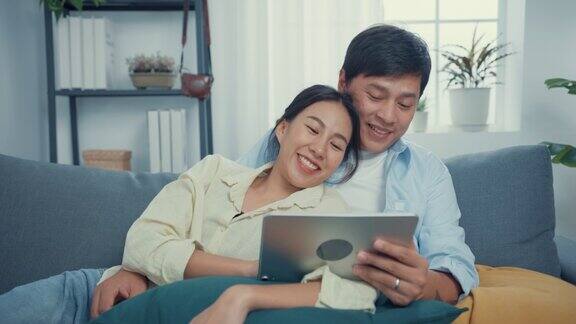 年轻的亚洲夫妇休闲地坐在沙发上使用平板电脑在线购物在家里的客厅观看在线娱乐电影