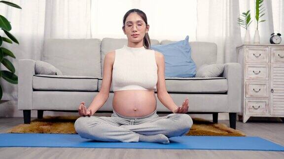 美丽的亚洲孕妇大肚子显示腹部坐在瑜伽姿势冥想在家里客厅的地毯上放松等待分娩