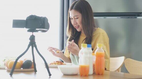 年轻的亚洲女性美食博主穿着围裙在她的美食频道录制吃意大利面番茄酱的视频一位女厨师一边吃着她的意大利面番茄一边在自家厨房用数码相机播报