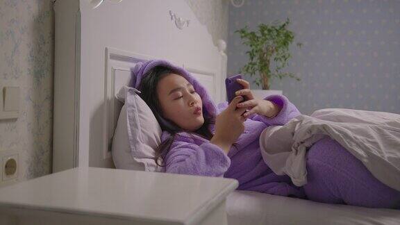 20岁亚洲女性躺在床上用手机上网穿着紫色睡衣的微笑女人在床上用手机上网
