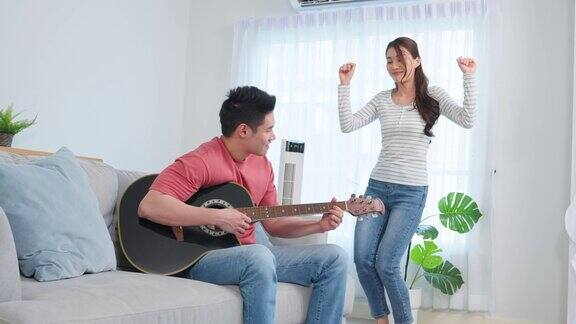 亚洲年轻情侣在家里的客厅里一起弹吉他迷人浪漫的新婚活跃浪漫的男士坐在沙发上弹着乐器与美丽的女友共舞