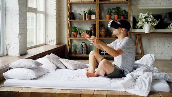 一名兴奋的年轻人坐在明亮公寓的双人床上用增强现实眼镜移动双手现代科技和快乐人的理念