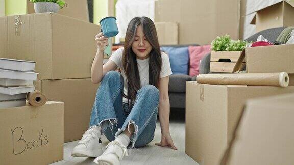 年轻的中国女子坐在新家的地板上用智能手机喝咖啡