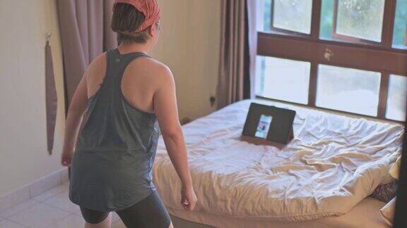 亚洲华人中年妇女戴头巾在家锻炼蹲下姿势锻炼在线学习健身教练在卧室通过智能手机在线互联网