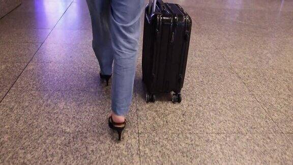 女人穿着牛仔裤长裤黑色鞋跟鞋带着黑色行李箱沿着机场航站楼走着看腿的底部
