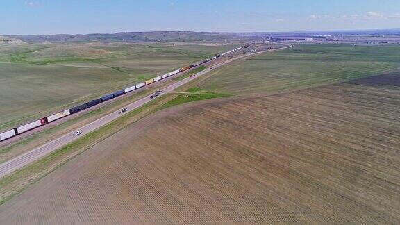早春时节一列货运火车经过北达科他州威利斯顿附近的田野汽车和卡车在铁路沿线的高速公路上行驶无人机视频与前进摄像头运动跟踪列车