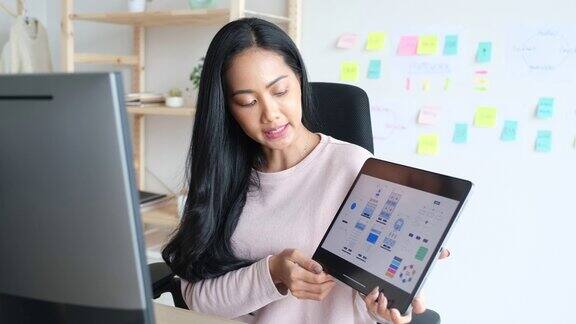 年轻的ui设计开发人员女程序员手持数字平板电脑屏幕上显示着移动应用程序原型同时与团队在家庭办公室进行虚拟视频会议在家工作