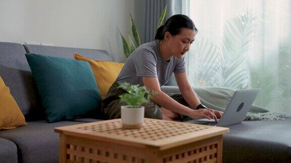 亚洲女性坐在家里的客厅里用笔记本电脑和键盘打字