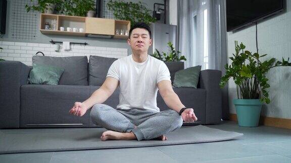 亚洲运动男子练习瑜伽坐在垫子上的莲花位置在家里的客厅冷静的男性在室内冥想和放松