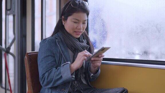 年轻快乐的亚洲女性在旅行期间坐在公共交通穿梭巴士的窗户边使用智能手机而外面下雨女性在欧洲度假旅行