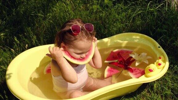 一个孩子坐在黄色的水盆里吃着西瓜水盆里还有水果片、玩具橡皮鸭和鲜花