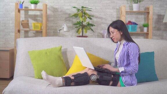 受伤后带着笔记本电脑的亚洲女性肖像膝盖弯曲矫正器膝盖支架躺在家里的沙发上