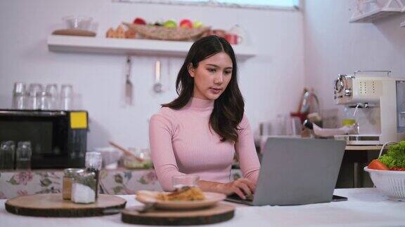 微笑的年轻亚洲女子坐在厨房里在家里的厨房里用笔记本电脑看搜索网上博客在家里享受周末