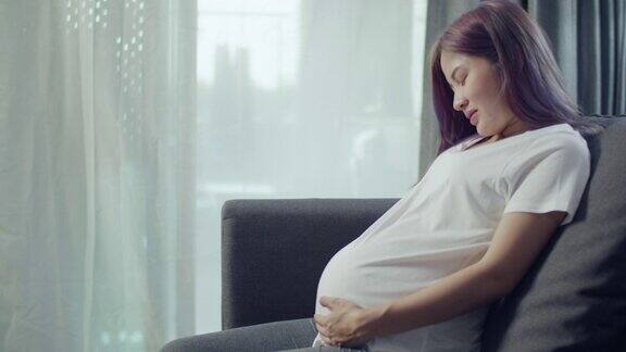 美丽的亚洲孕妇抚摸肚子而坐在沙发上的客厅在家里