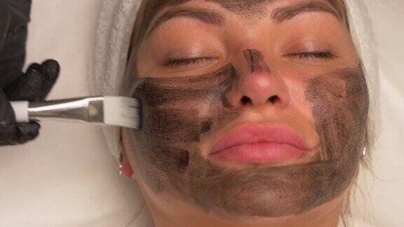 美容师在戴着黑色手套的漂亮女人脸上涂上黑色面膜漂亮的女人在水疗中心做面部手术