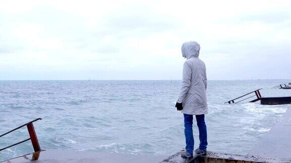 一个穿着白色夹克的女人站在海边的海滩上看着暴风雨的海浪后视图