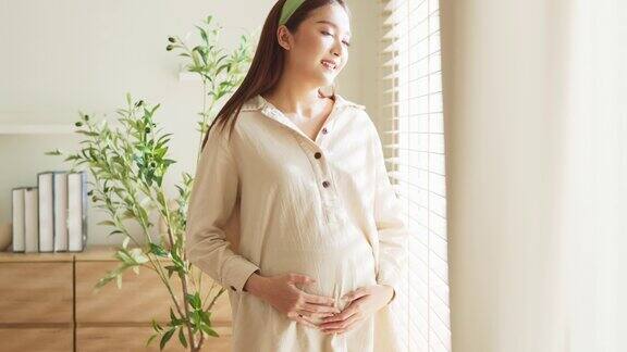 亚洲成年孕妇妈妈站在窗边用手温柔地抚摸着宝宝在家里抚摸着她的肚子