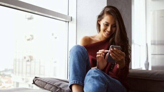 一个快乐的黑发女人穿着毛衣和牛仔裤坐在窗边用着智能手机