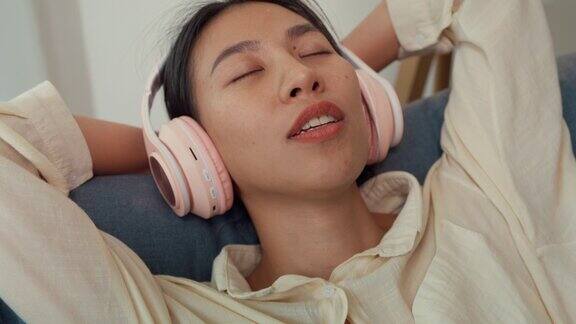 特写年轻的亚洲女性闭着眼睛躺在沙发上休息戴着耳机听音乐在音频流媒体广播上唱喜欢的歌曲在家在线播客