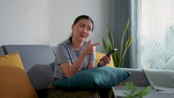 亚洲女性坐在家里的沙发上看着相机使用智能手机感到困惑