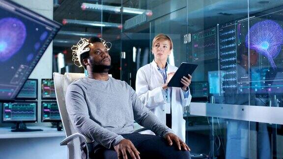 戴着脑电波扫描耳机的人坐在椅子上而科学家用平板电脑监督过程在现代大脑研究实验室监测显示脑电图阅读和大脑模型