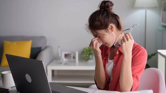年轻的亚洲妇女坐在眼睛闭从疲惫和疲倦在工作在家庭办公室使用笔记本电脑劳累过度的亚洲女学生工作人员在电脑工作后因疲劳而按摩眼睛和太阳穴