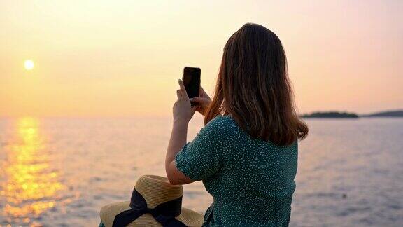 年轻女子坐在岩石上用智能手机在平静的海面上拍摄日落