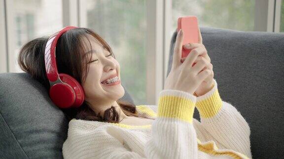 亚洲少女躺在沙发上戴着耳机听音乐用智能手机在社交媒体、休闲概念、社交距离、物联网上交流