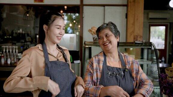 年长的母亲带着亚洲女儿站在收银台前取咖啡这是自己的面包店站在他们的围裙口袋在口袋里举手做心
