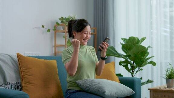 亚洲女人坐在家里客厅的沙发上看着智能手机开心兴奋地赢得了比赛