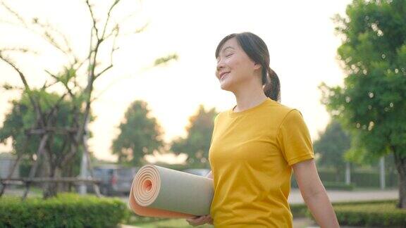 亚洲妇女开心地微笑着穿着运动服抱着卷好的瑜伽垫去公园做瑜伽练习