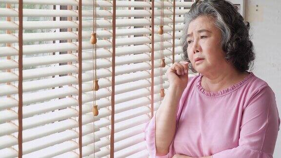 退休妇女独自在家一个悲伤的老妇人独自站在窗前想着一些事情:自然光线中的悲伤和沮丧