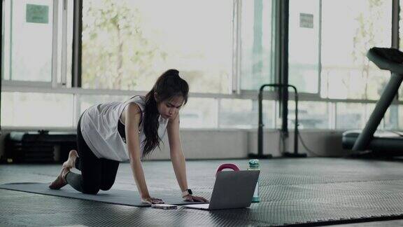年轻的亚洲妇女在笔记本电脑上学习在健身房做瑜伽冥想