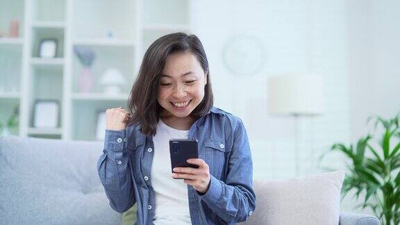 快乐的亚洲女性坐在家里客厅的沙发上用智能手机看新闻庆祝成功