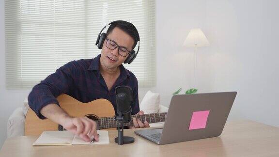 亚洲人戴着耳机弹吉他唱歌用笔记本电脑和笔记本电脑在线学习写歌录音用电脑录制音乐程序音乐家