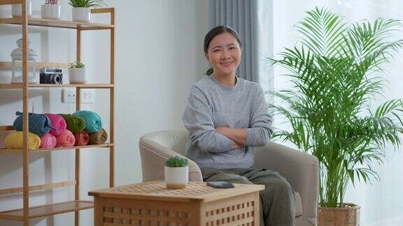 亚洲女性在家里坐在扶手椅上看着相机和挥动手指感到无聊