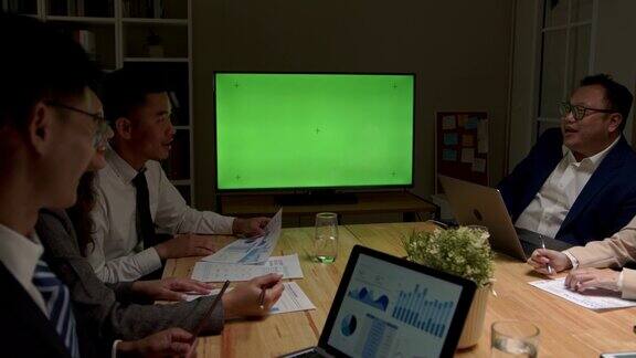 一群成功的亚洲商务人士在会议室与绿色屏幕色度键电视在夜间办公室