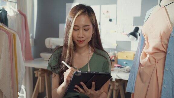 开朗的专业亚洲女性时装设计师随意使用数码平板电脑在她的商店创造新的系列女裁缝书房在写字板上画草图微笑着看着工作室里的相机