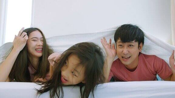 快乐迷人的亚洲家庭父亲母亲和女儿女孩微笑着笑着一起度过时光躺在卧室的毯子下父母和可爱的孩子看着镜头